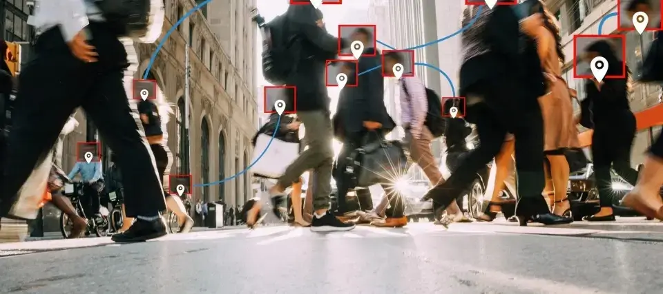 Street-level view of people walking across a busy city intersection, overlaid with facial recognition boxes and location icons, symbolizing digital surveillance, tracking, and data monitoring in public urban spaces.