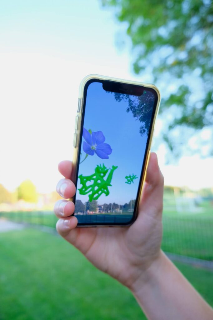 A hand holds a smartphone displaying a virtual blue flower and green shapes against a clear sky. Nearby, a sunlit park with trees adds a serene atmosphere.