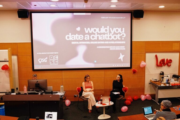 A conference room with valentine's decor and a presentation titled "Would You Date a Chatbot?" Two women sit on stage, audience members visible.