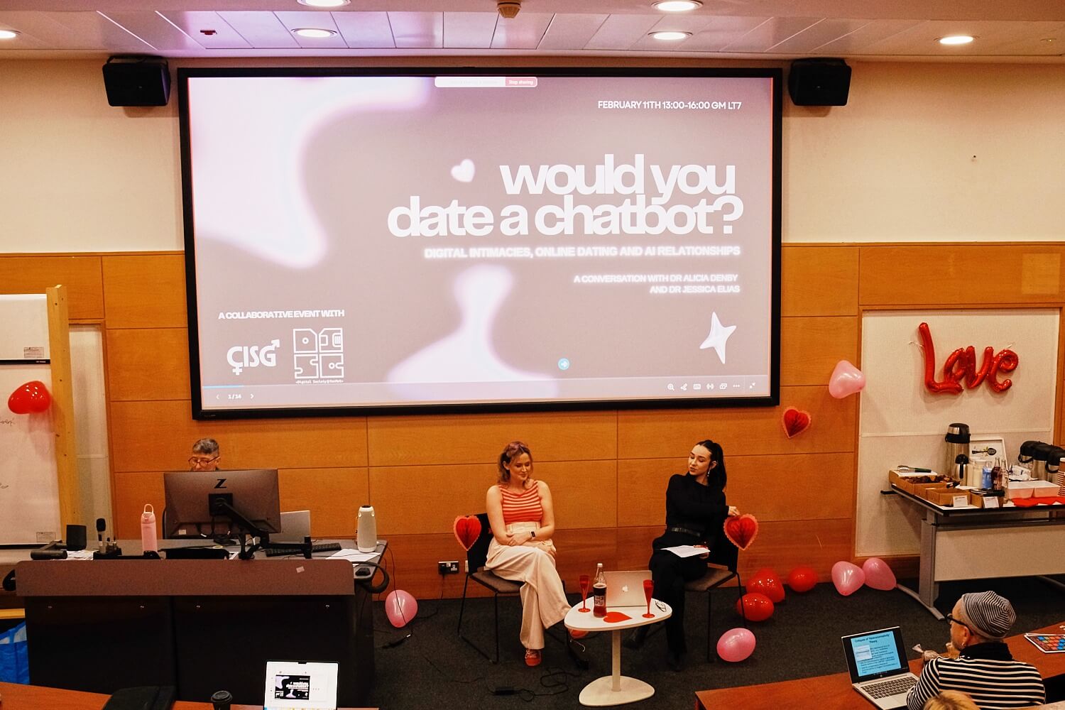 A conference room with valentine's decor and a presentation titled "Would You Date a Chatbot?" Two women sit on stage, audience members visible.