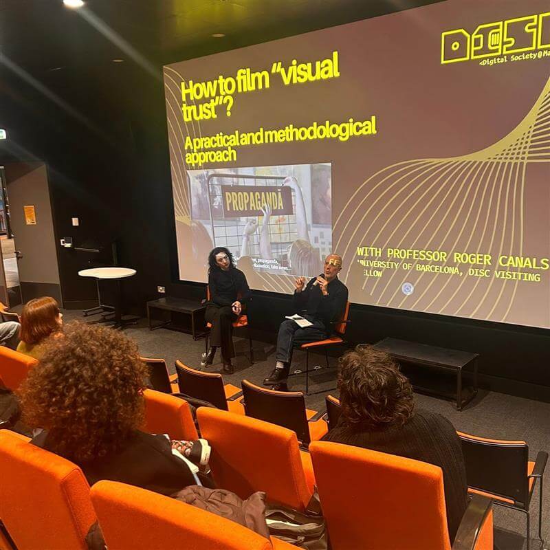 Two speakers sit on chairs in front of an audience in an auditorium. Behind them, a large screen displays a presentation title: "How to film 'visual trust'? A practical and methodological approach".