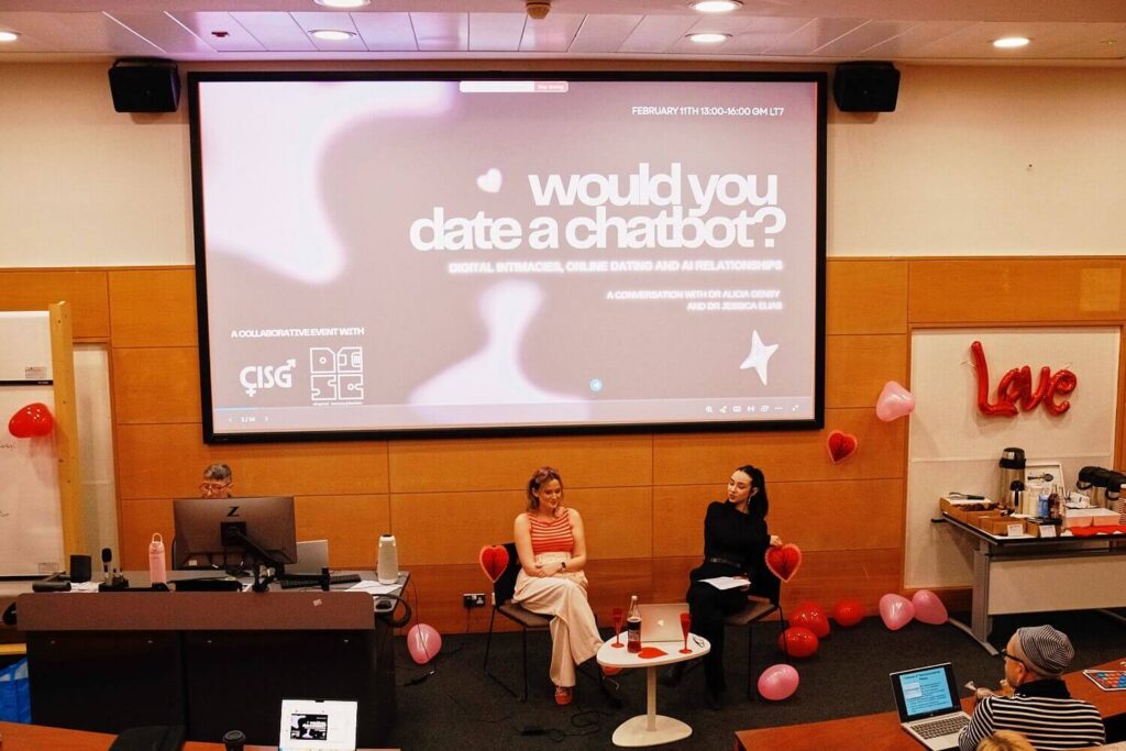 Two women seated at a table, engaged in conversation, with a screen displaying information in the background.